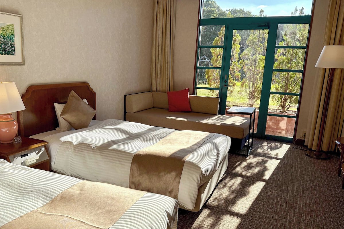 Ein Hotelzimmer mit zwei Betten, einem Sofa, einem Tisch und einem Fenster mit Blick auf Bäume.