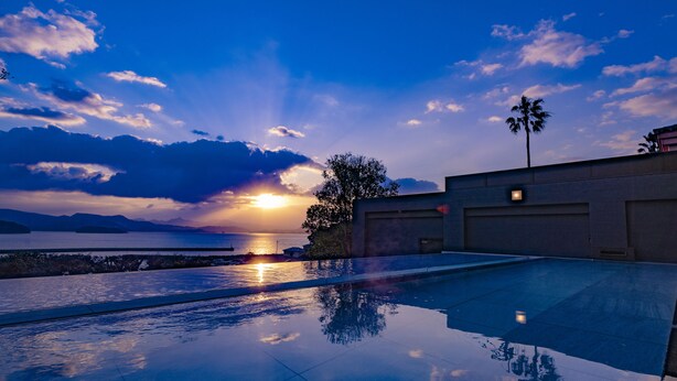 A swimming pool with a sunset view, a palm tree, and a building with a garage. - Ooedo Onsen Monogatari Premium Saikaibashi (Sasebo)