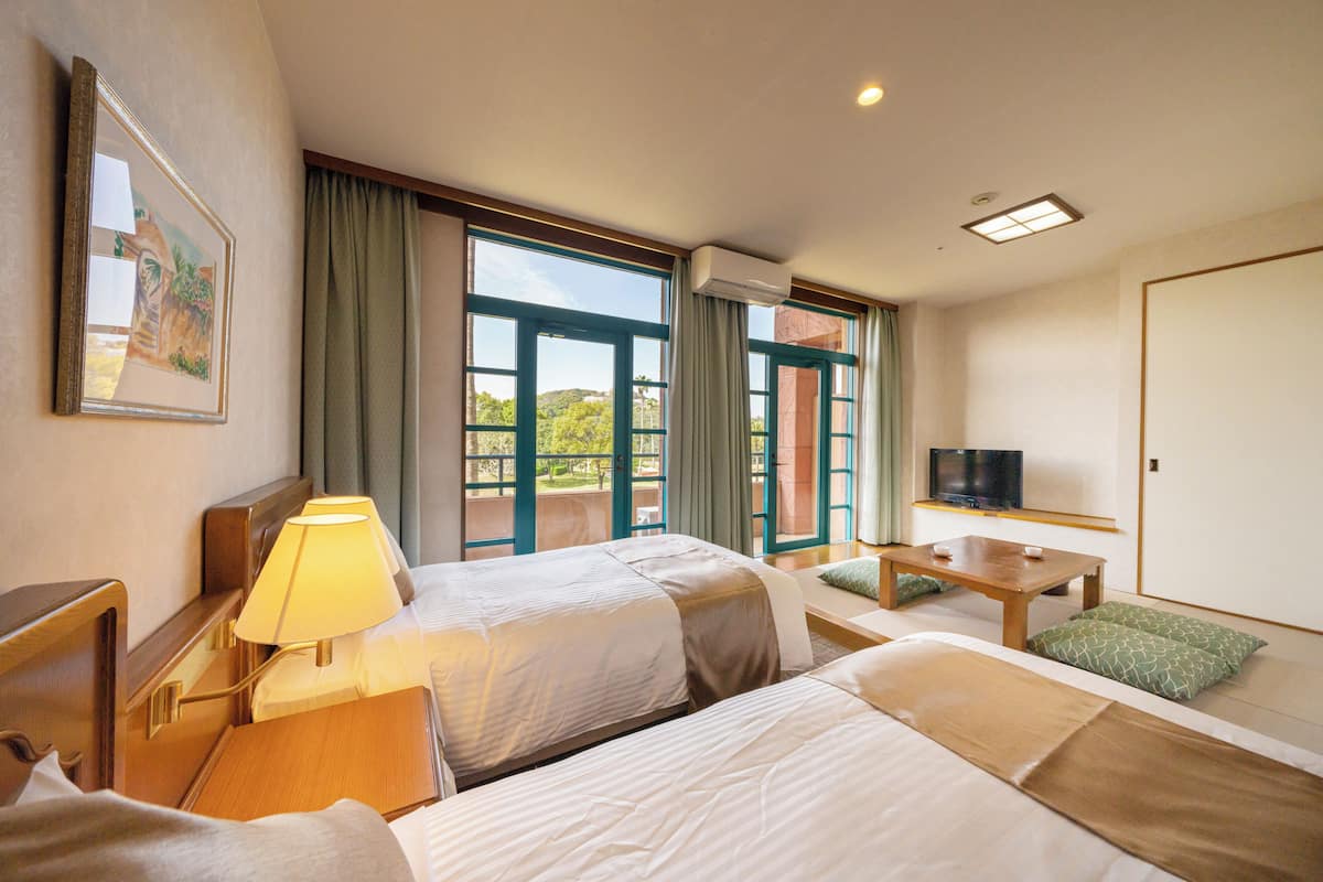 Ein Hotelzimmer mit zwei Betten, einem Schreibtisch, einem Fernseher und großen Fenstern mit Vorhängen.