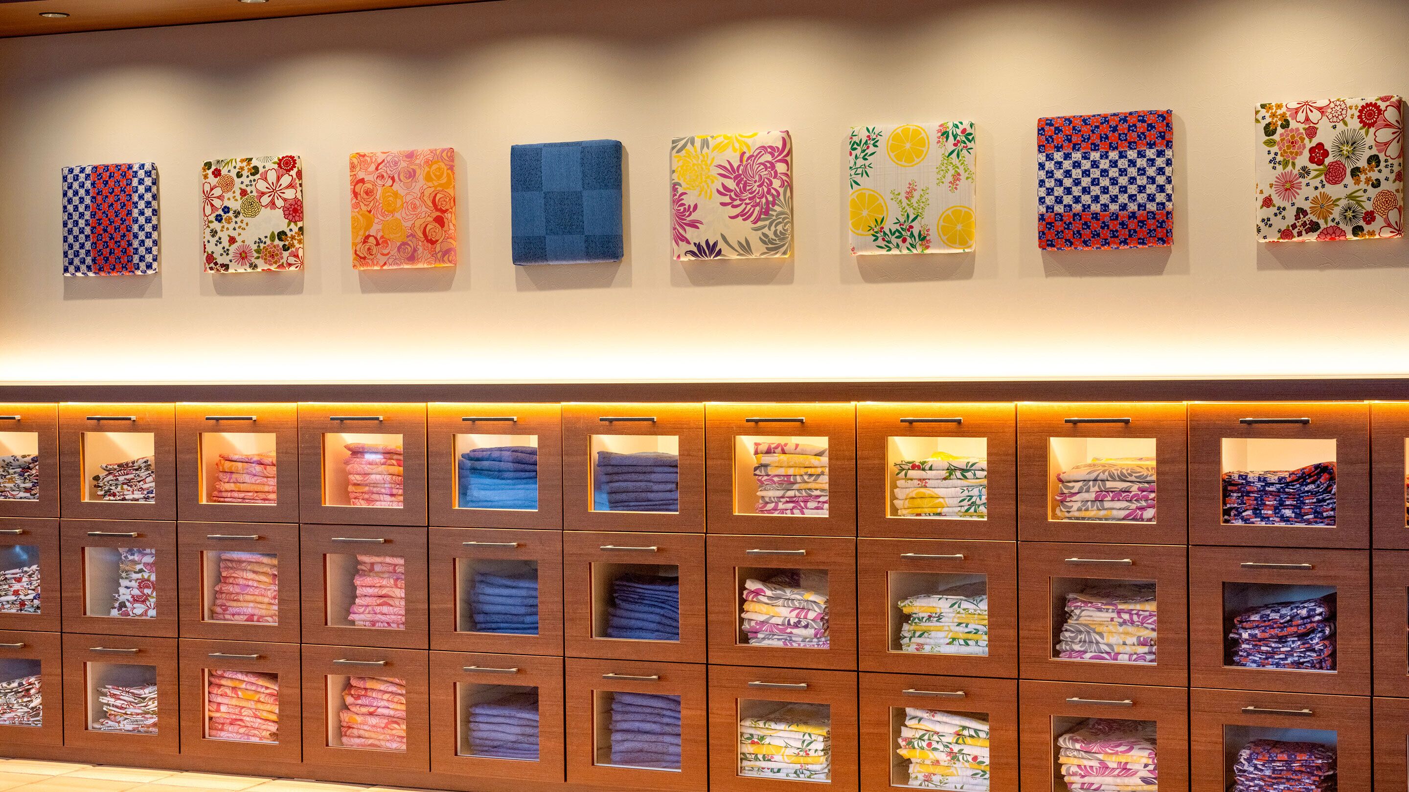 Pared de hotel con una exhibición de toallas dobladas cuidadosamente en varios colores y diseños, y una serie de obras de arte enmarcadas arriba.