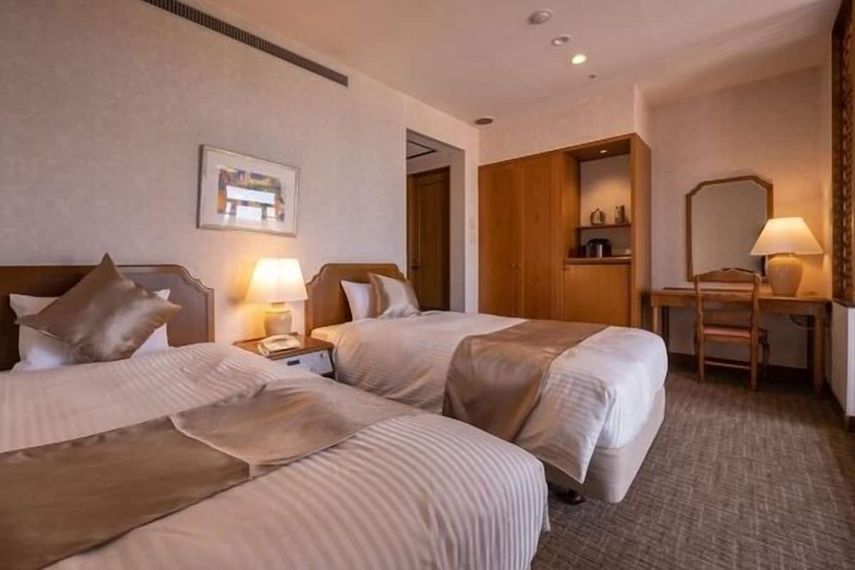Ein Hotelzimmer mit zwei Betten, einem Schreibtisch, einem Stuhl, einer Lampe und einem gerahmten Bild an der Wand.