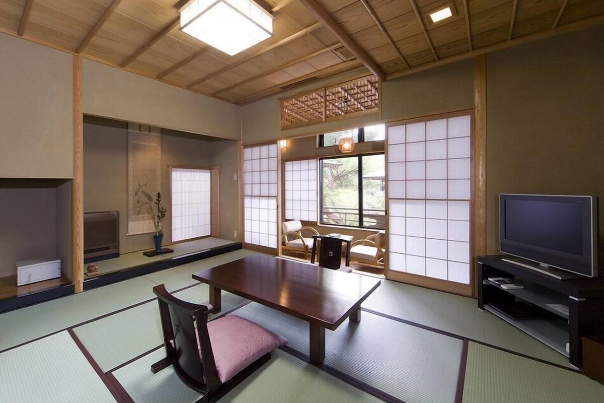 Una habitación japonesa tradicional con suelo de tatami, una mesa baja, sillas, un televisor y puertas correderas.