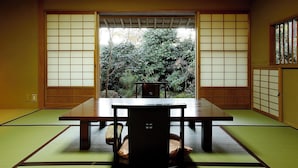 Una habitación con esteras tatami, una mesa baja, una silla, puertas correderas amplias y vistas a un jardín.