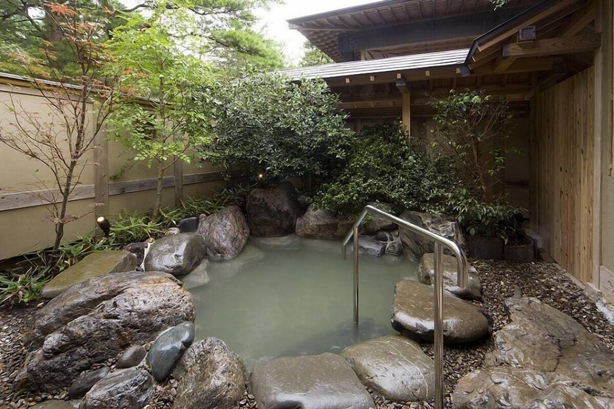 Un onsen japonés tradicional con estructura de madera, una fuente termal revestida de roca y un sendero de piedra.