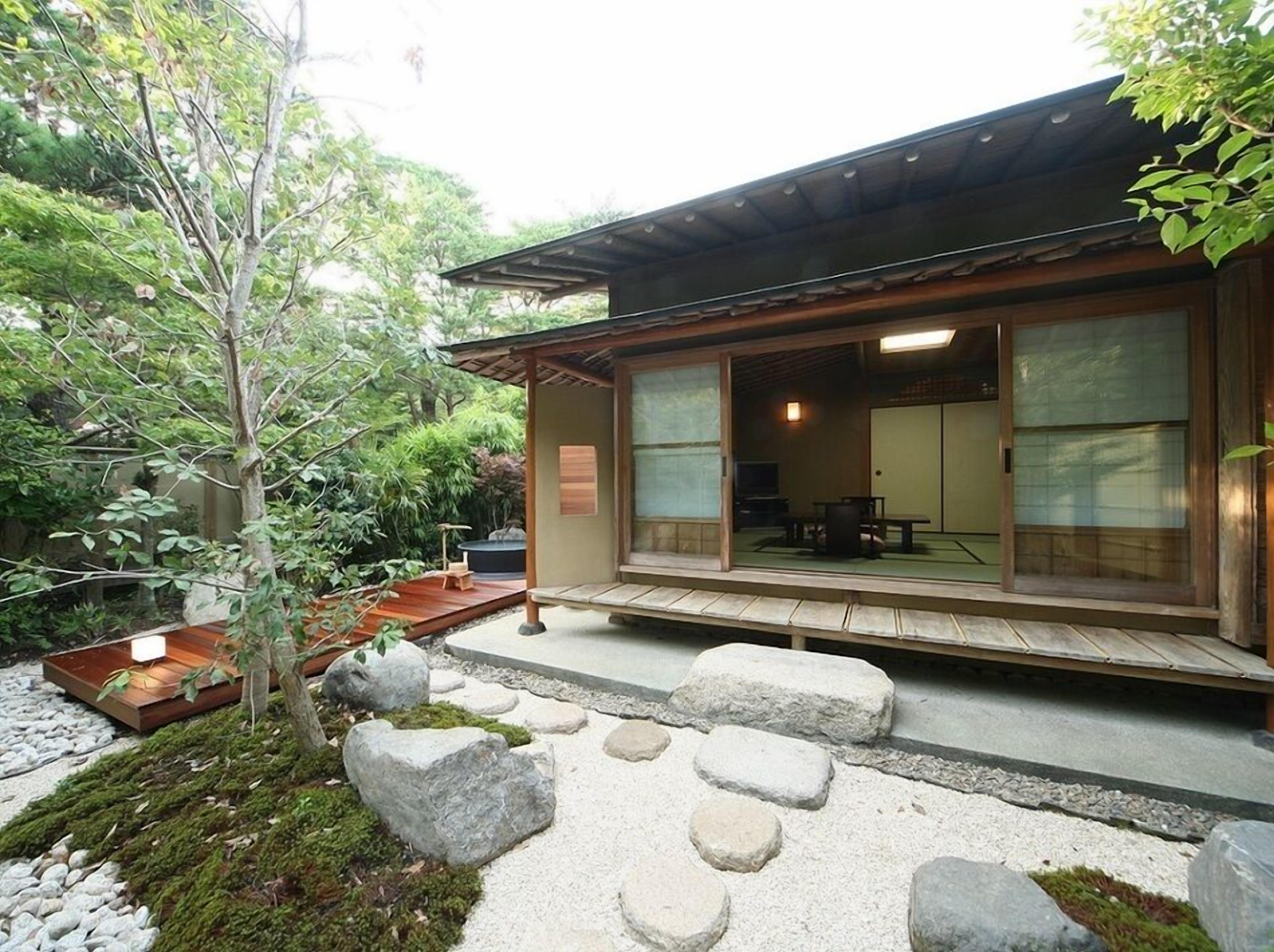 Um edifício tradicional em estilo japonês, com fachada de madeira, caminho de pedra e um pequeno jardim com pedras e uma árvore.