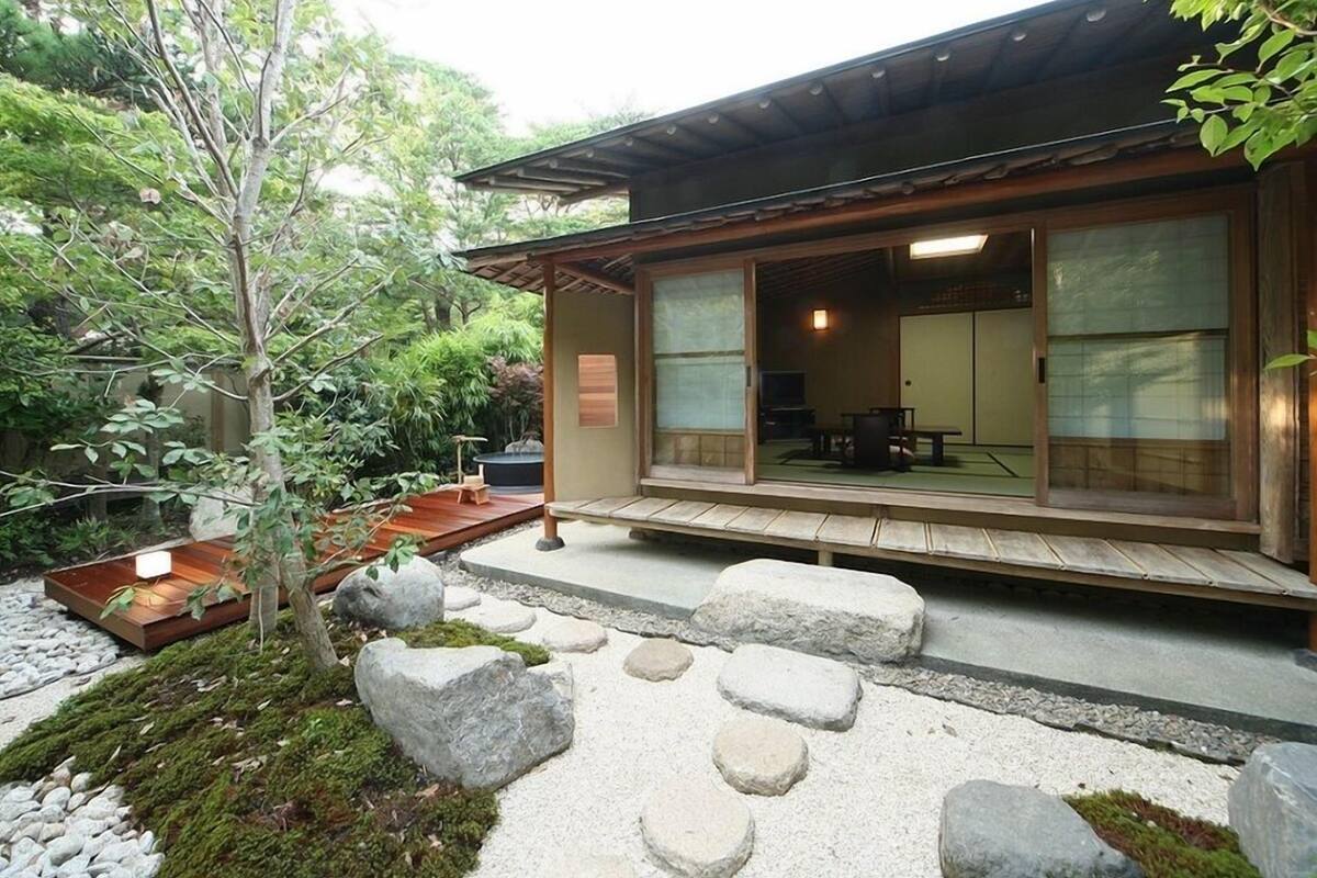 Edificio tradicional japonés con fachada de madera, camino de piedra y pequeño jardín con rocas y un árbol.