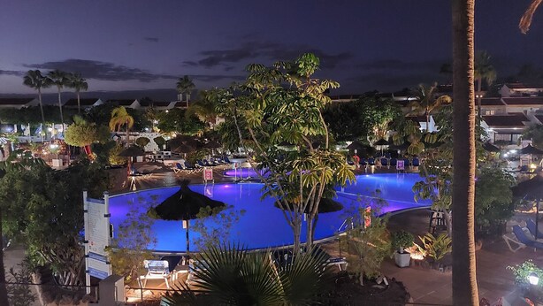 Vista noturna de uma área de piscina em resort, com espreguiçadeiras e guarda-sóis, cercada por palmeiras e céu limpo.