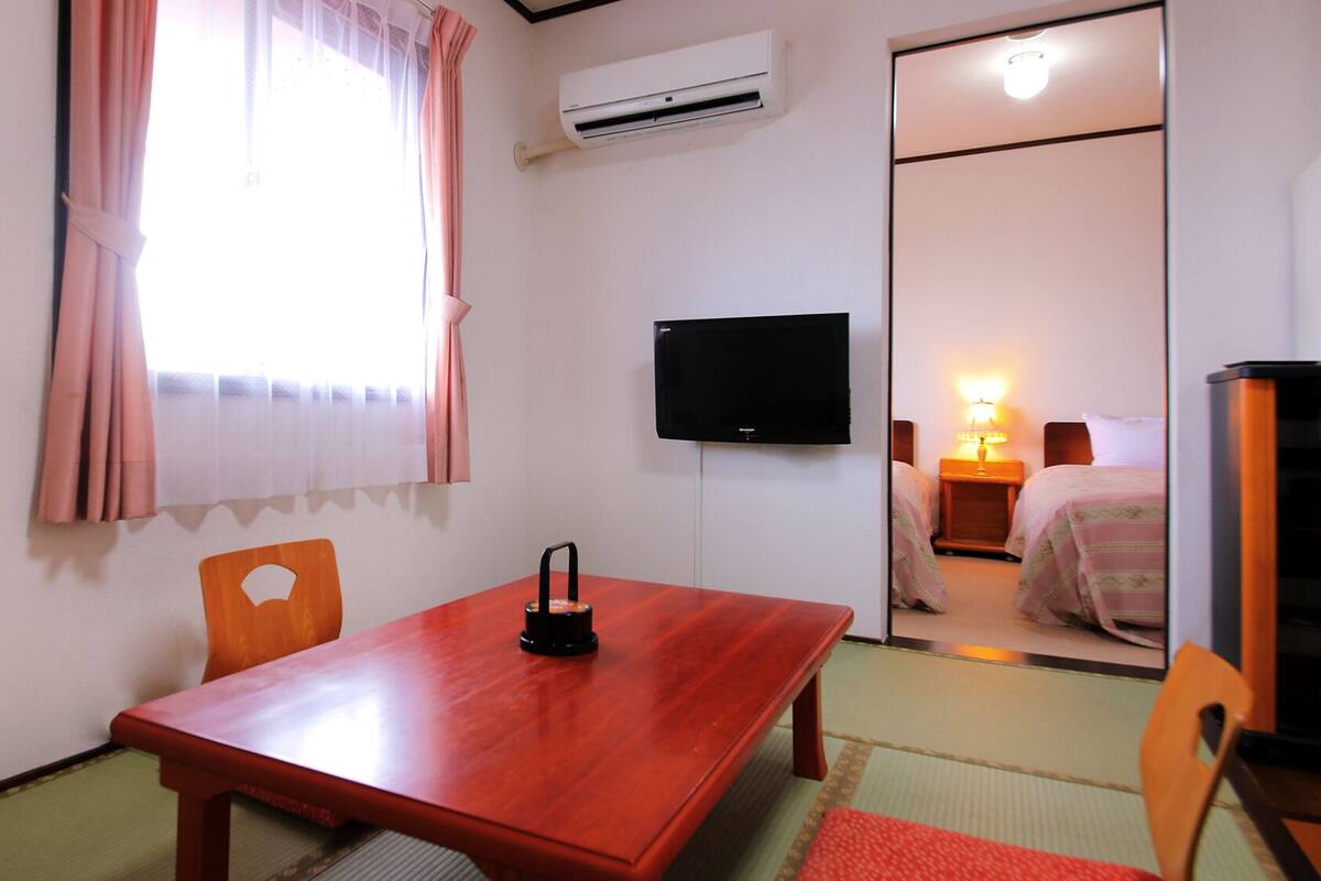 A room with a wooden table, a TV mounted on the wall, an air conditioner, and a bed with a nightstand.