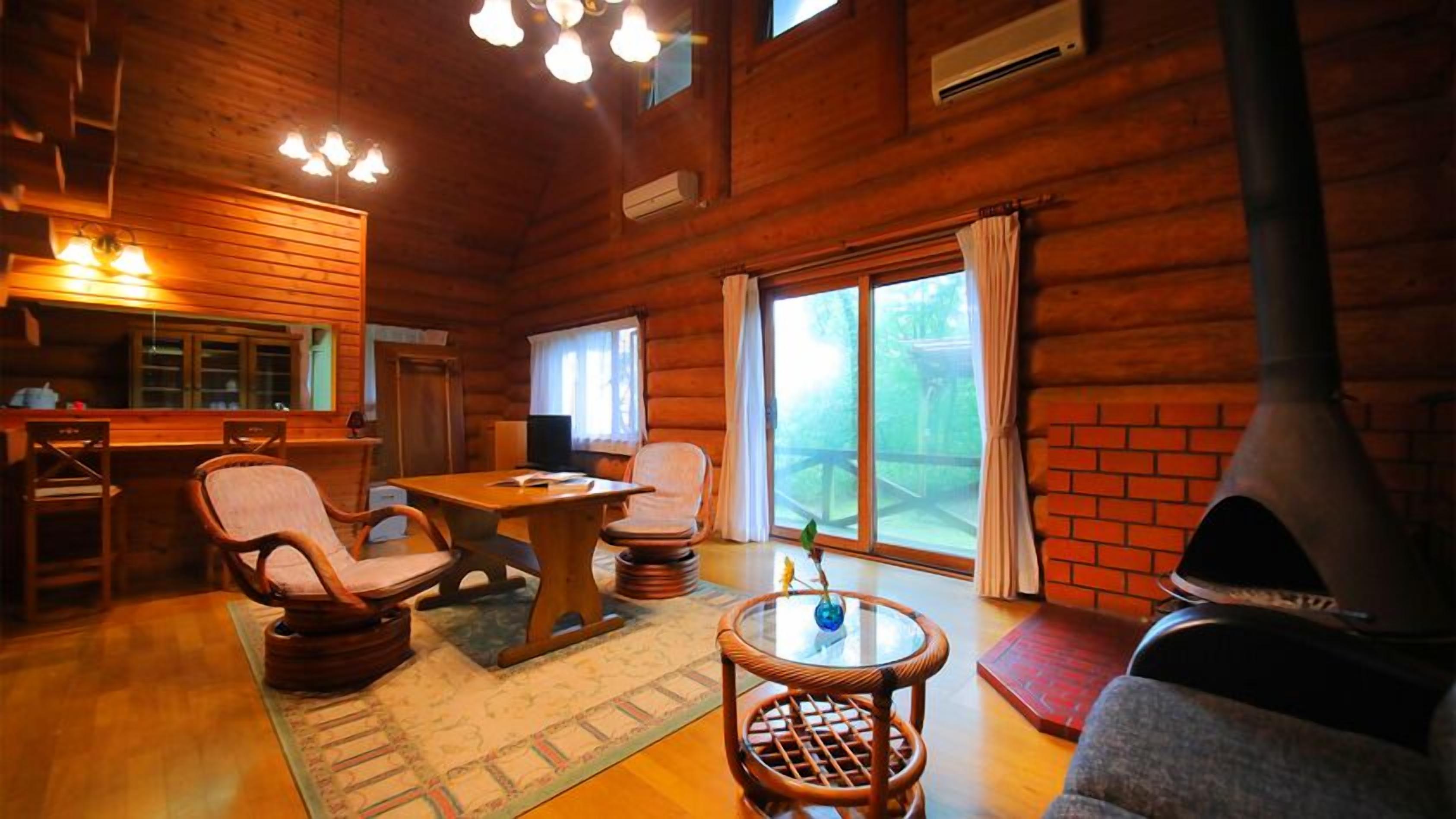 A wooden cabin interior with a dining table, chairs, a small round table with a vase, a fireplace, and a large window with curtains.