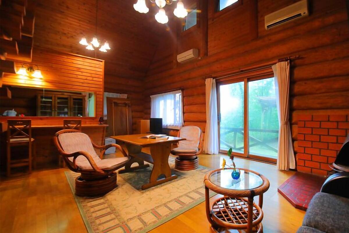 A wooden cabin interior with a dining table, chairs, a small round table with a vase, a fireplace, and a large window with curtains.
