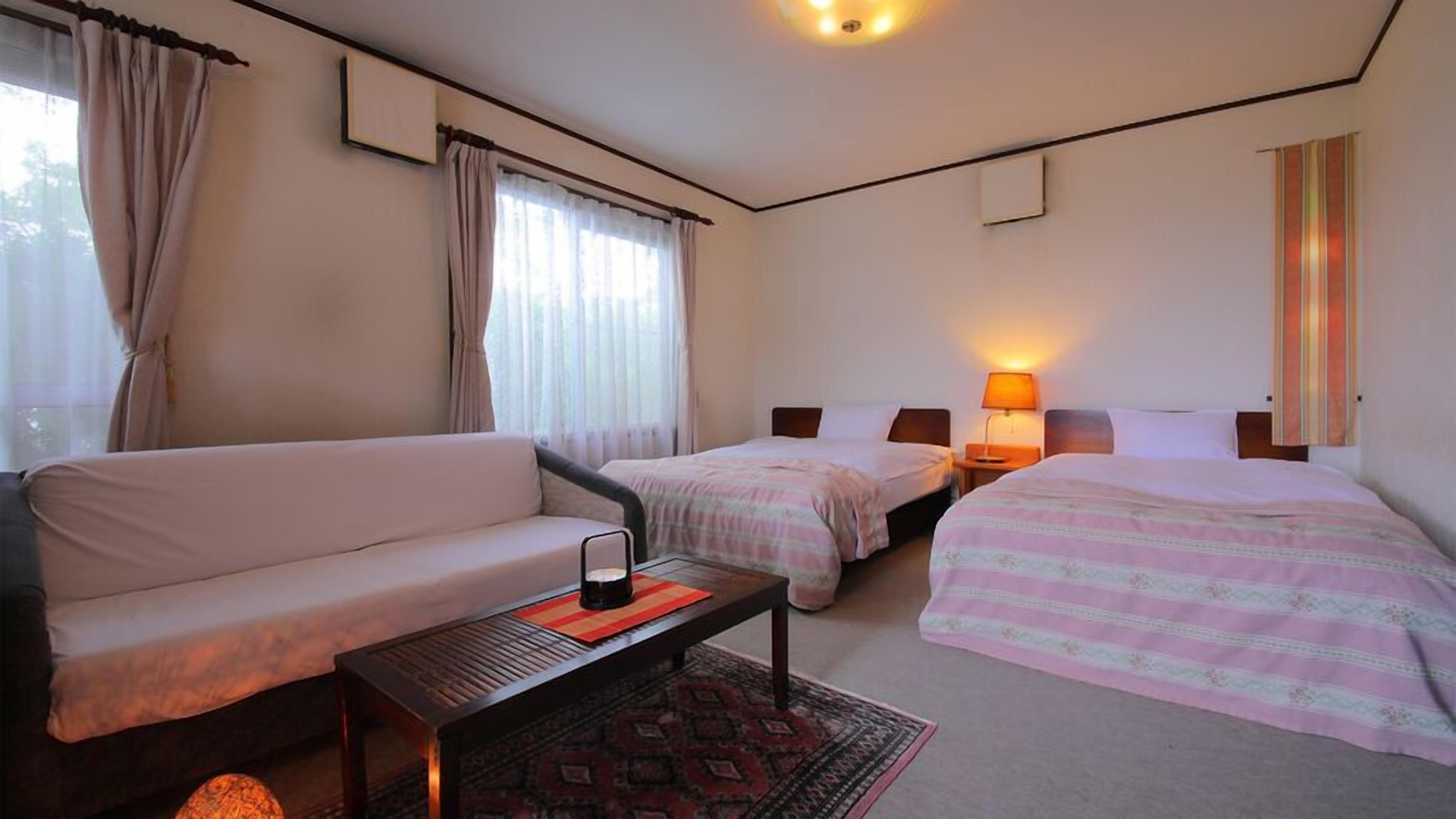 A hotel room with a sofa, two single beds, a small table, and a lamp.