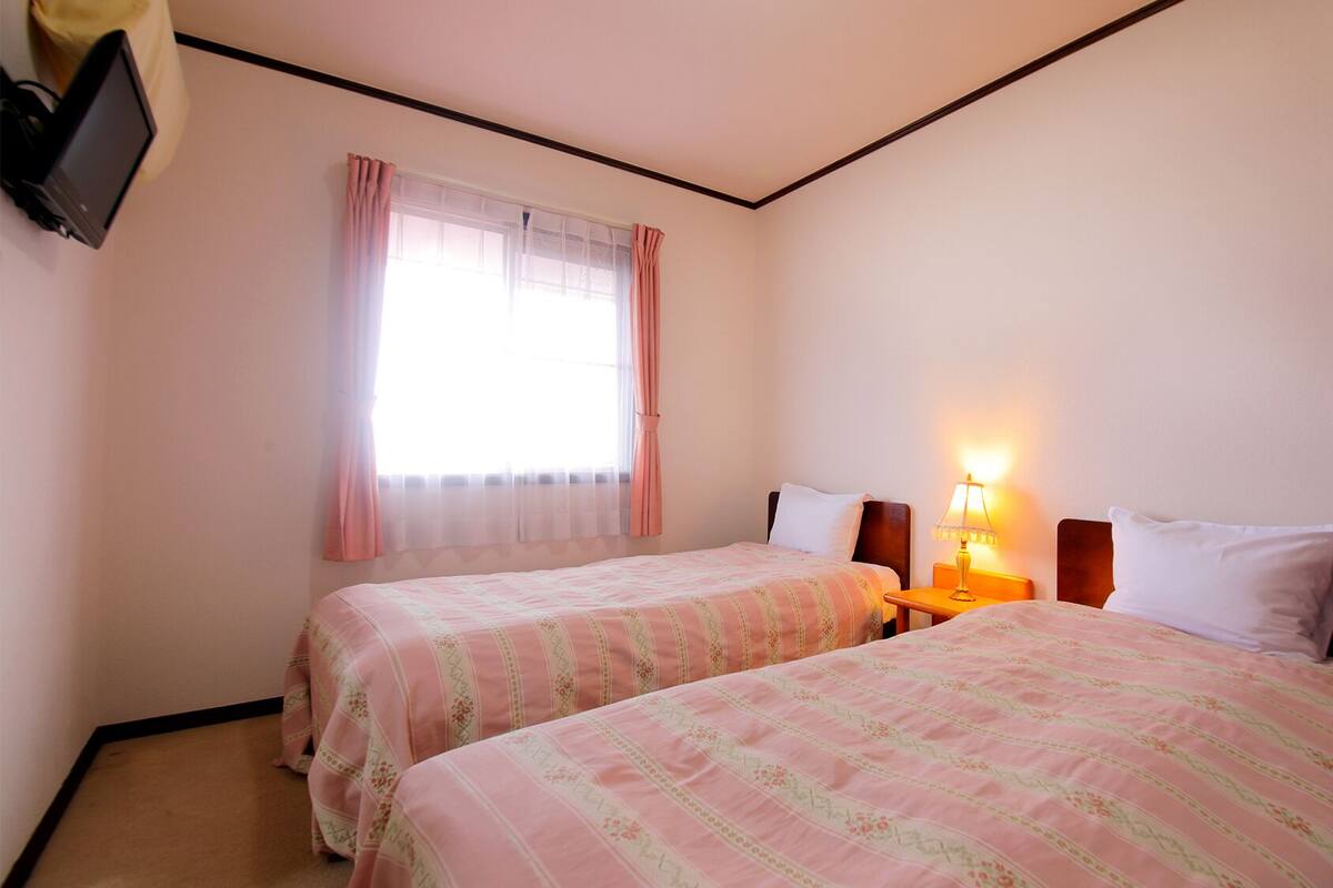 A hotel room with two beds, a window with curtains, a bedside lamp, and a television on the wall.