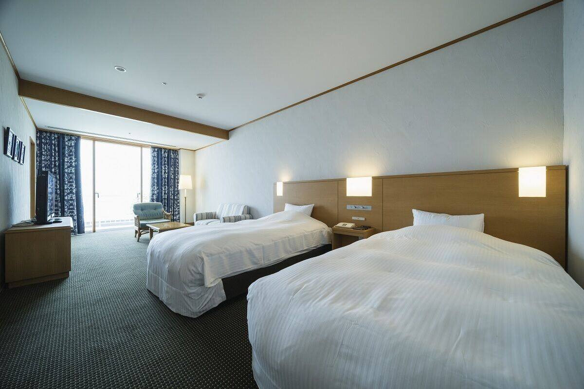 A hotel room with two beds, a desk, a chair, and a view of the outdoors.