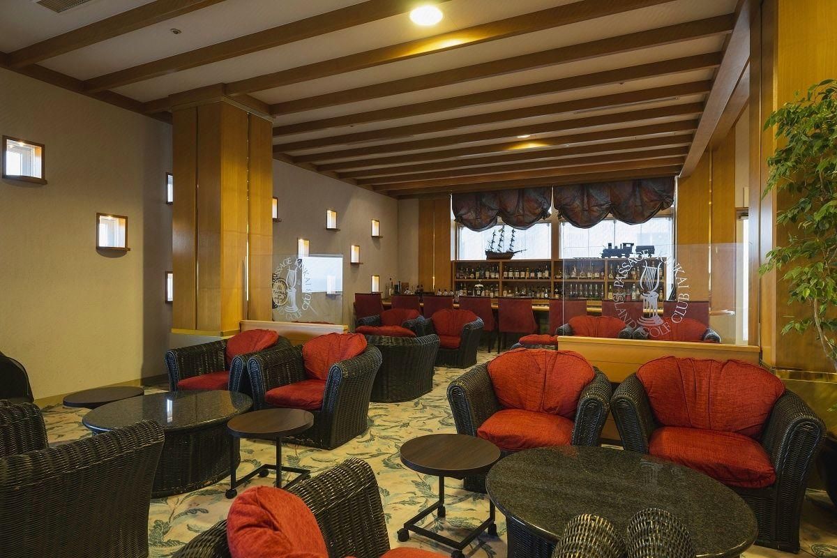 A lounge area with wicker furniture, a bar in the background, and a view of a ship through the window.
