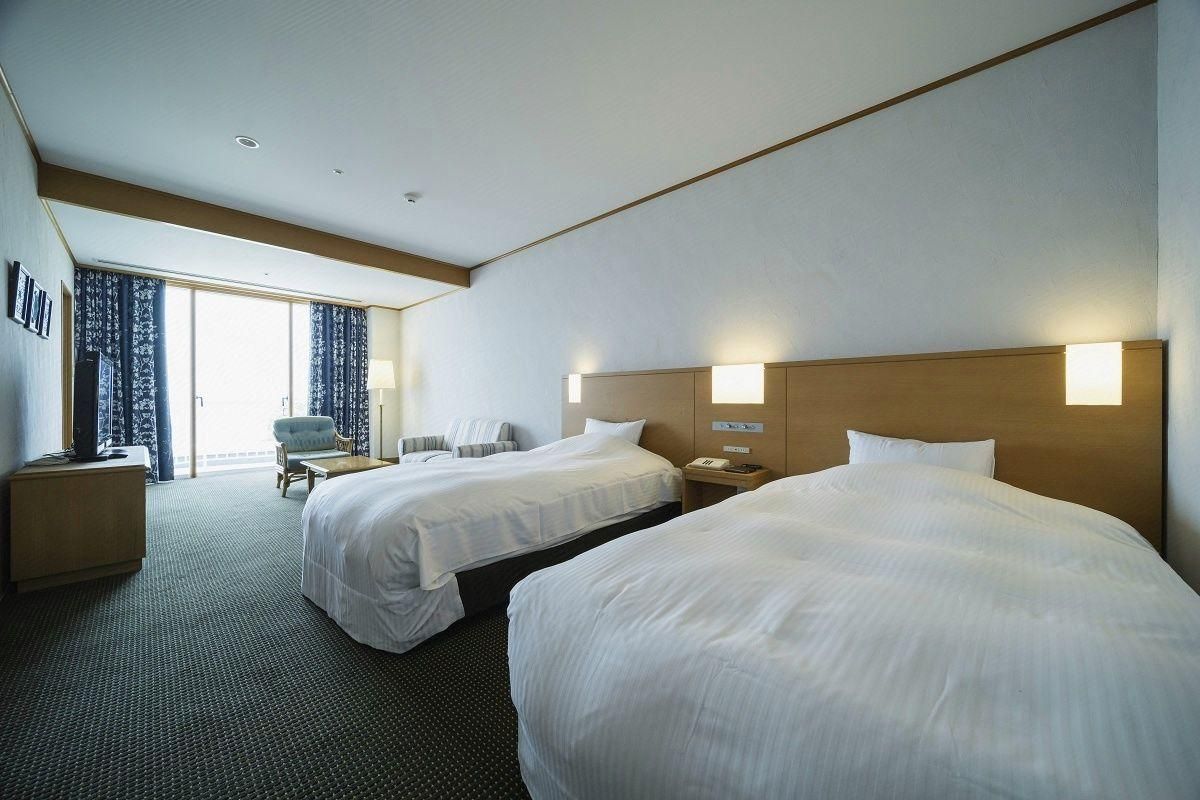 A hotel room with two beds, a desk, a chair, and a view of the outdoors.