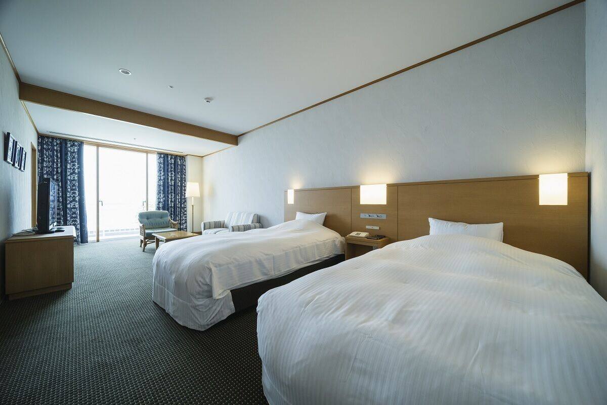 A hotel room with two beds, a desk, a chair, and a view of the outdoors.