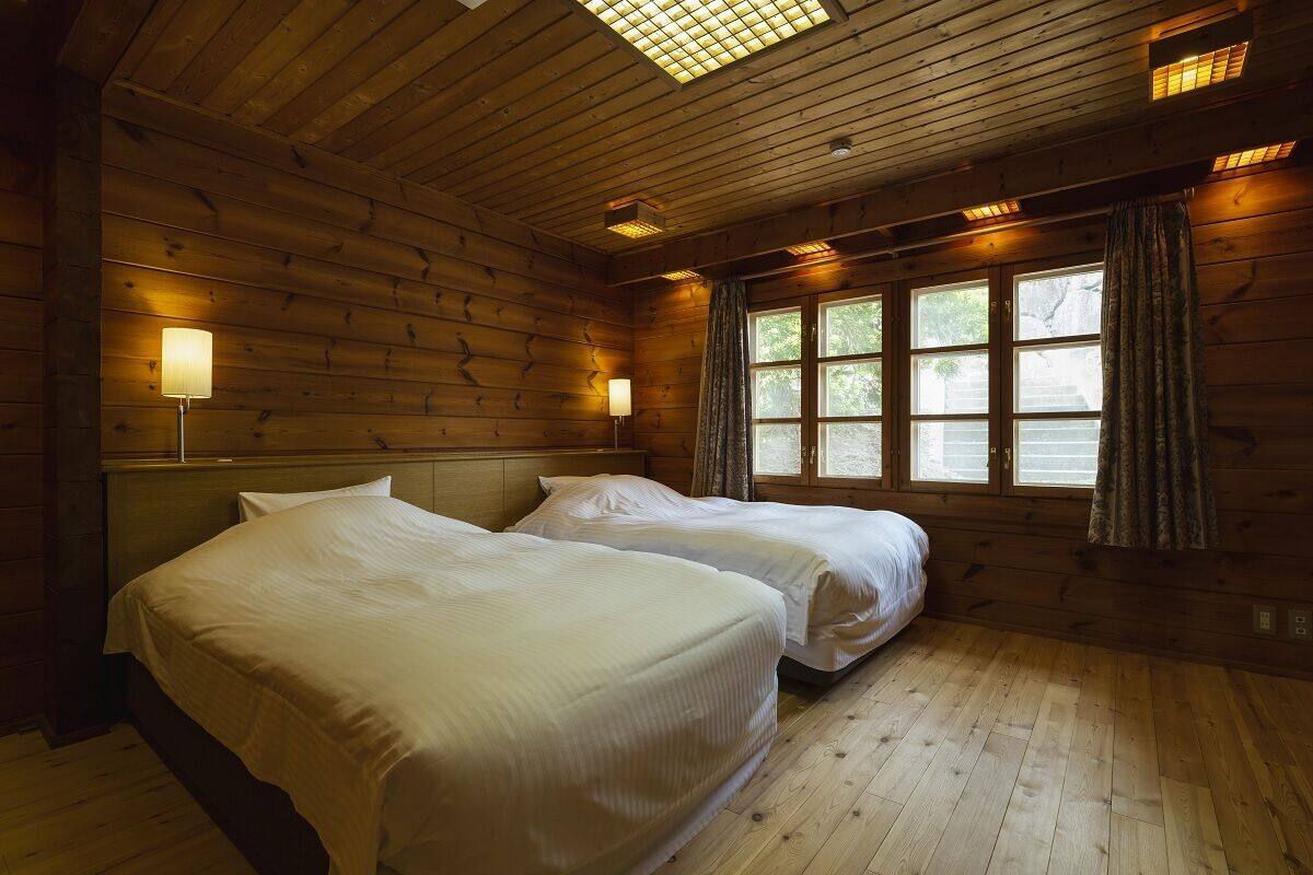 A room with wooden walls, two beds, a window with curtains, and a ceiling light fixture.