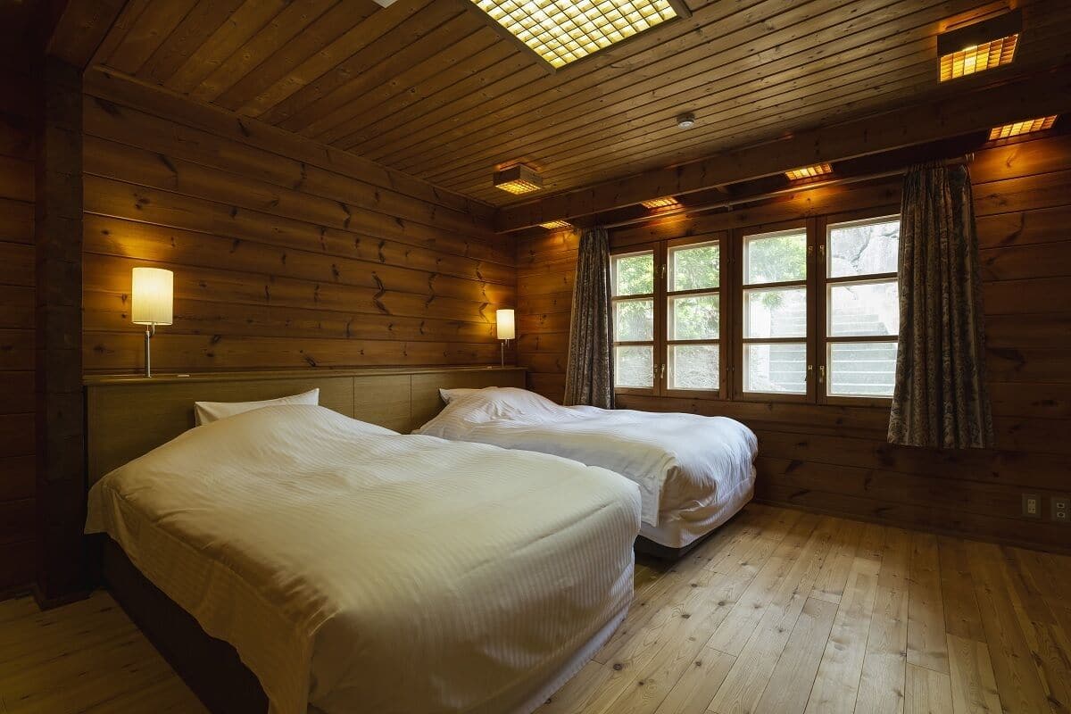 Una habitación con paredes de madera, dos camas, una ventana con cortinas y una lámpara en el techo.