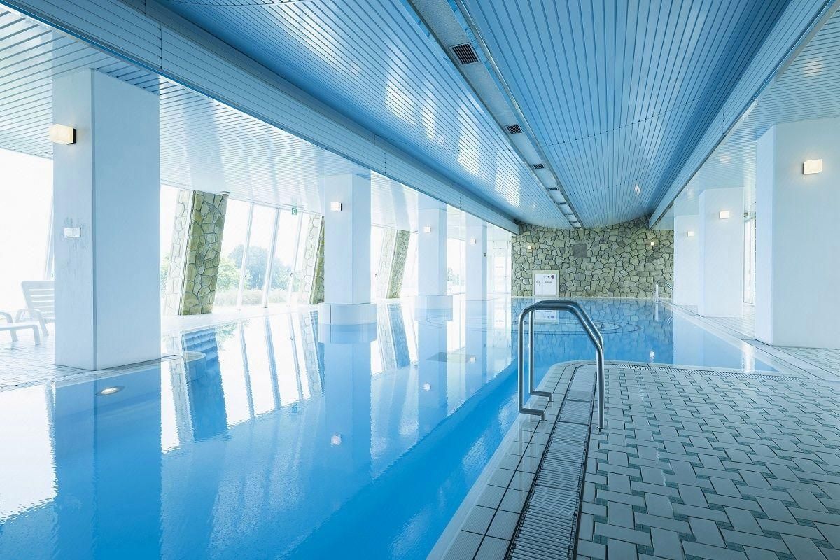 Ein großes Hallenbad mit klarem, blauem Wasser, umgeben von weißen Wänden und einer Wand mit Steinverkleidung.
