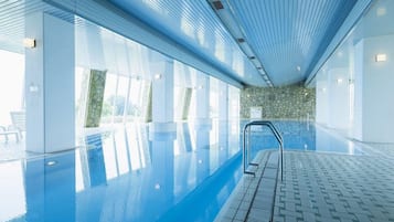 Ein großes Hallenbad mit klarem, blauem Wasser, umgeben von weißen Wänden und einer Wand mit Steinverkleidung.