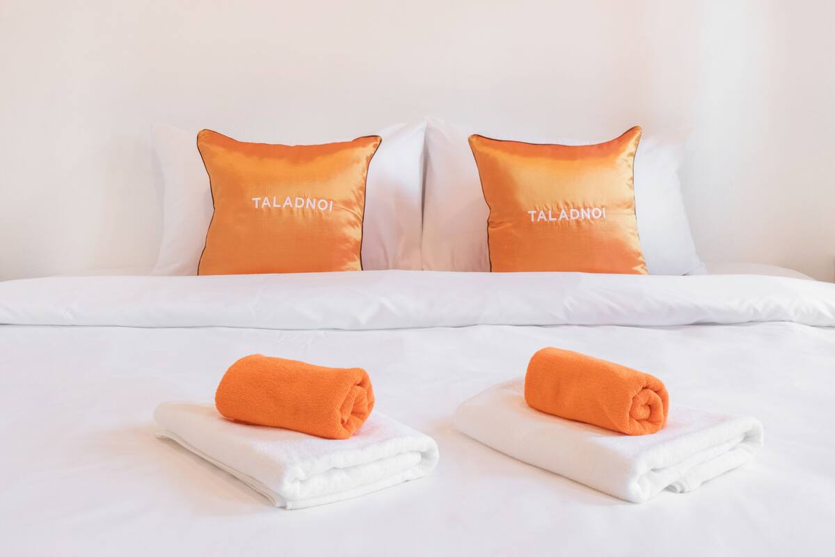 A hotel room with a neatly made bed, two pillows with the brand name 'TALADNOI', and two folded towels on the bed.