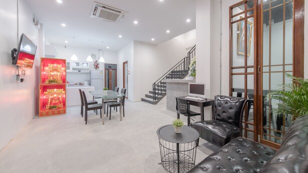 A spacious living area with a sofa, a dining table, and chairs. There is a kitchen in the background with a refrigerator and a microwave. A staircase leads to an upper floor. - Tiny Taladnoi Hostel (Bangkok)