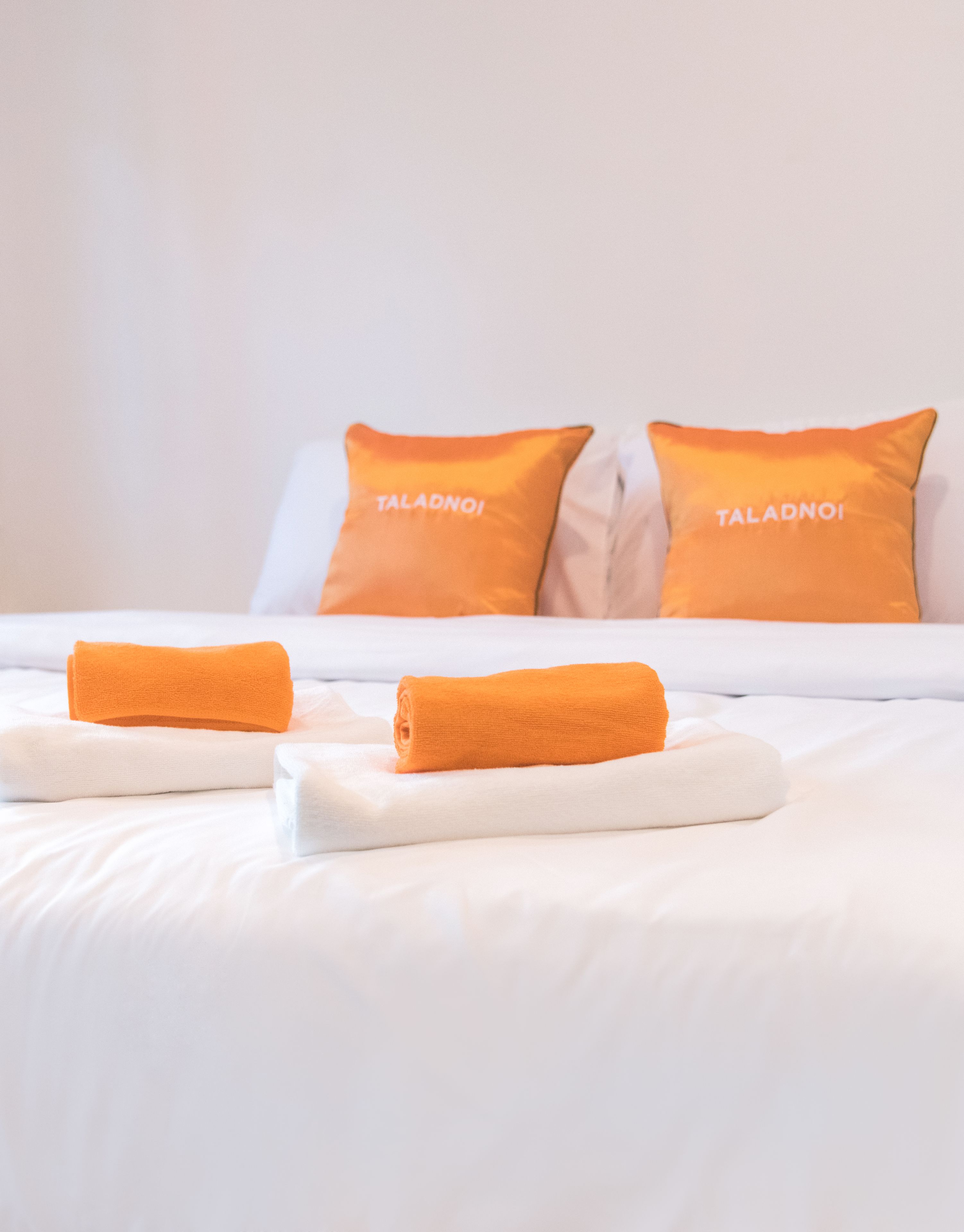 A hotel room with white bedding, two orange pillows with the text "TALADNOI", and two rolled orange towels on the bed.
