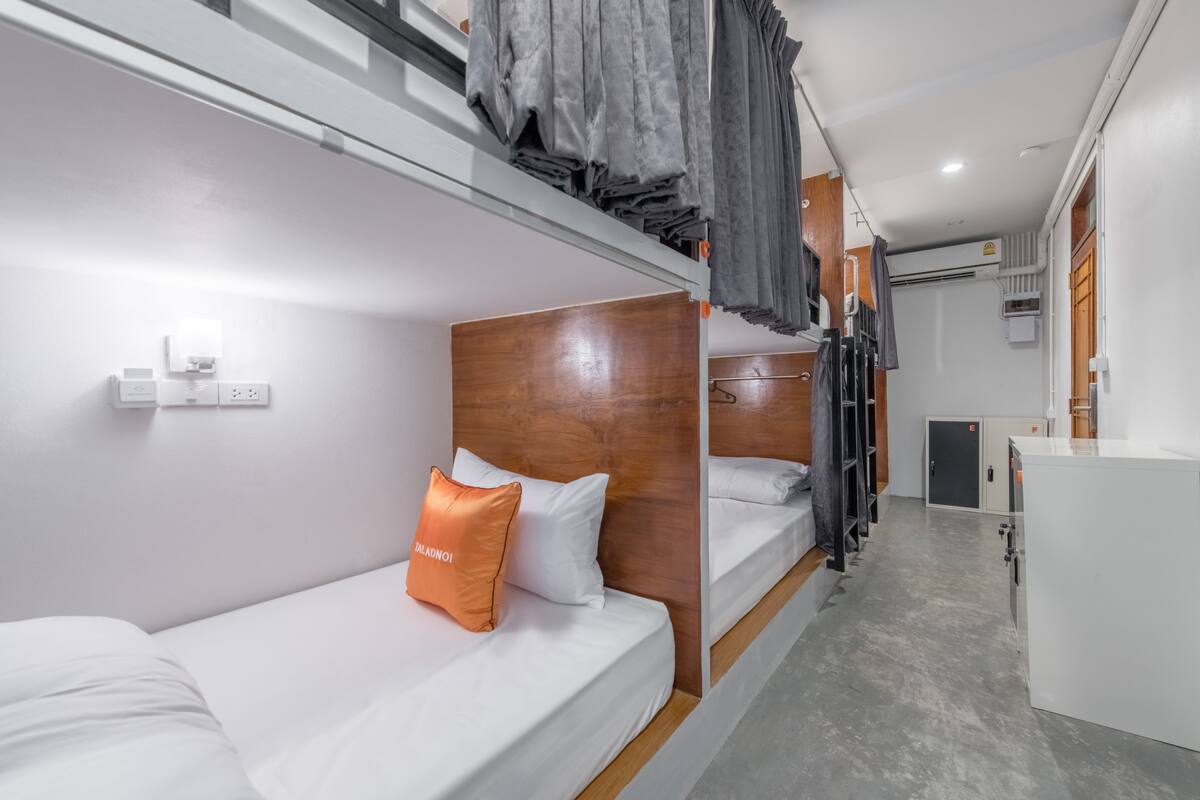 A bunk bed room with a wooden headboard, a white mattress, and an orange pillow. There is a small white cabinet and a grey curtain in the background.