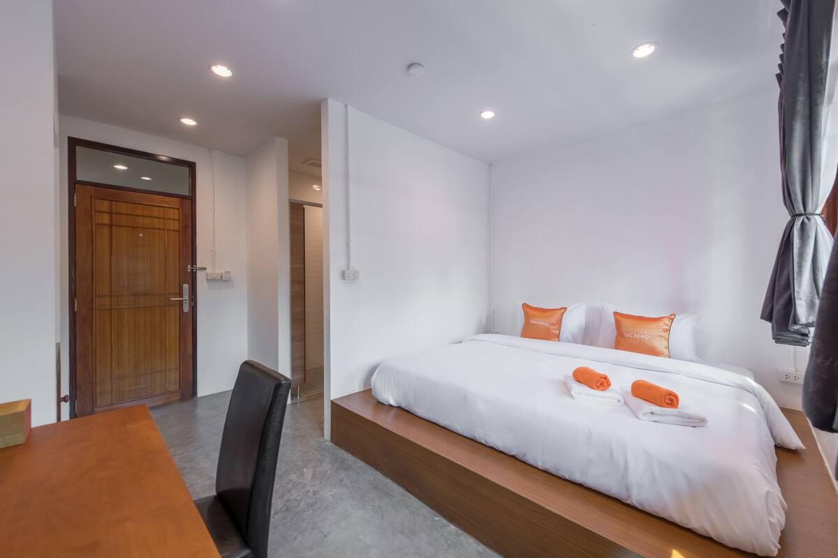 A modern hotel room with a wooden door, a bed with white bedding and orange pillows, a small table, and a chair.