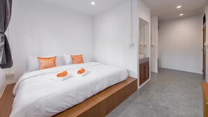 A modern hotel room with a large bed, white bedding, orange pillows, a wooden bench, a small bathroom, and a grey floor. - Tiny Taladnoi Hostel (Bangkok)