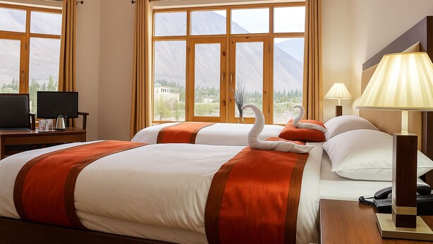 A hotel room with two beds, a desk with a computer, a lamp, and a view of mountains through large windows. - Hotel Grand Nubra (Leh)