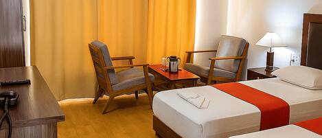 A hotel room with two single beds, a desk, two chairs, a table with a coffee pot, and a lamp.