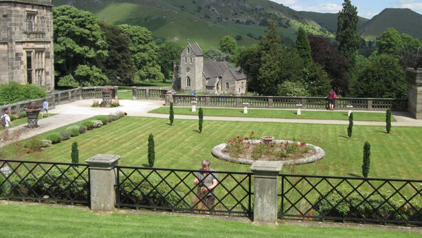A historic building with a stone facade, a formal garden with a central circular flower bed, and a view of rolling hills in the background.