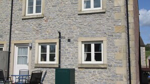 A stone building with three windows and a door, a small patio with chairs, and a green utility box.