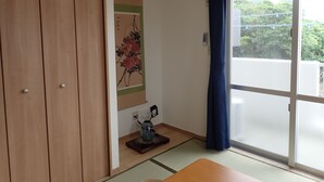 A room with a sliding glass door, a wooden wardrobe, a wall-mounted air conditioner, a small table with a vase, and a framed artwork.