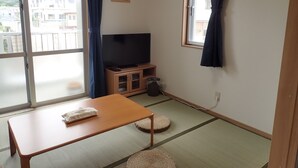 A room with tatami mats, a television on a wooden stand, and a sliding door with a view of buildings outside.
