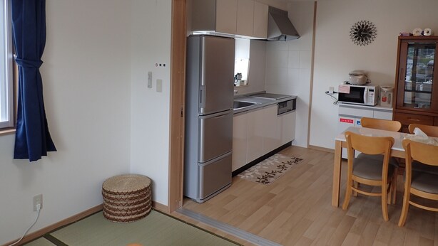 A compact kitchen with a stainless steel refrigerator, a microwave, and a sink. There is a dining table with chairs and a small cabinet with dishes.