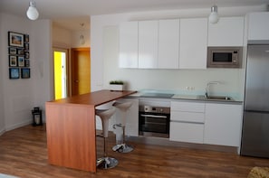 Una cucina moderna con mobili bianchi, un frigorifero in acciaio inossidabile, un forno a microonde e un forno integrato. È presente un'isola in legno con sgabelli da bar e una pianta in vaso sul piano di lavoro.