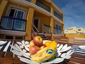 Un tavolo con una ciotola di frutta contenente mele, banane e arance, sullo sfondo di un edificio giallo con balconi e finestre.