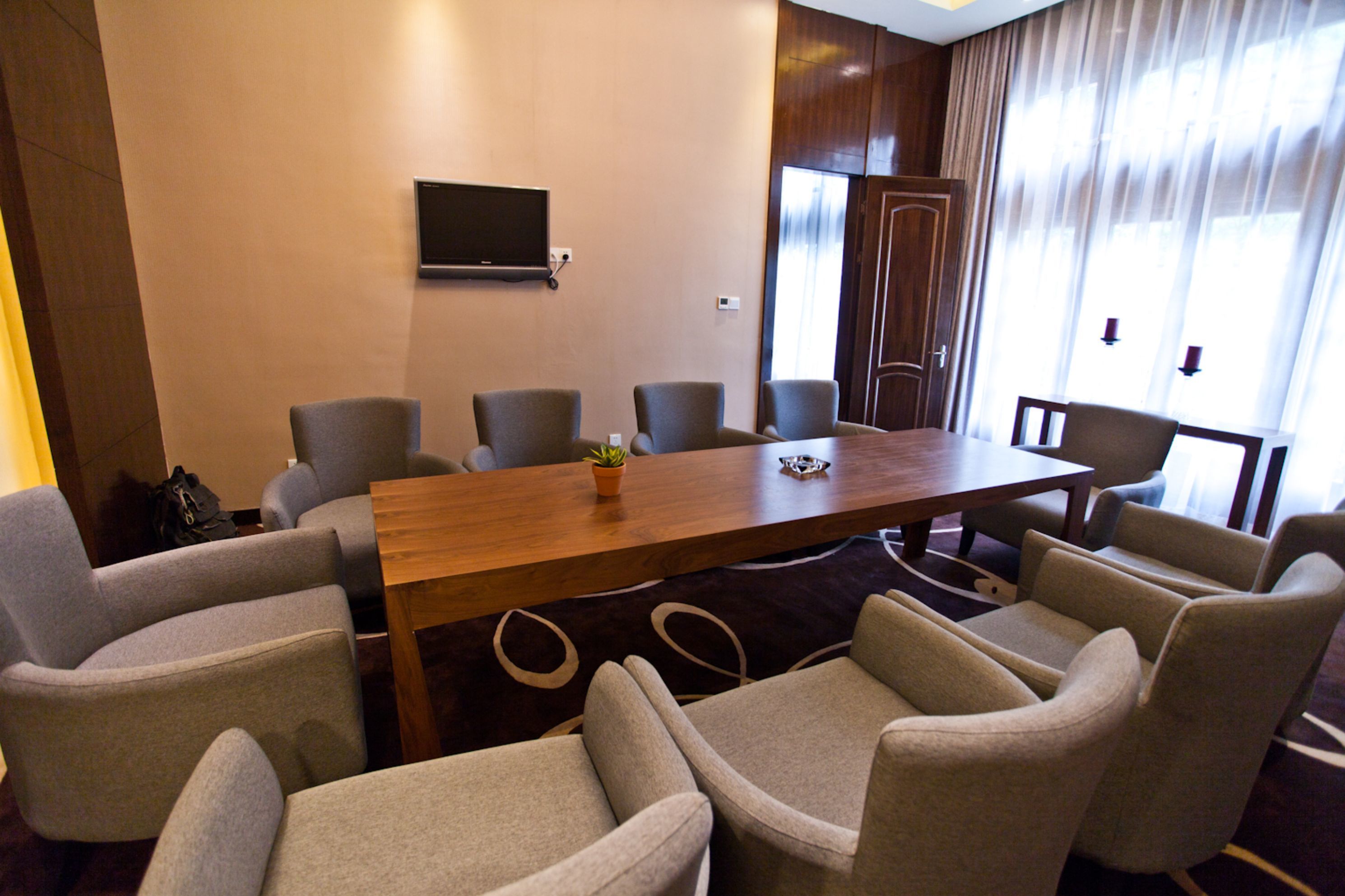 A conference room with a long table, six chairs, a television on the wall, and a large window with curtains.