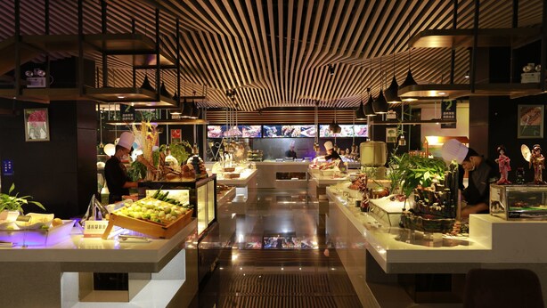 A modern restaurant with a central serving area, multiple food display counters, and a variety of plants and decorative items. - Jasmine International Hotel (Changsha)