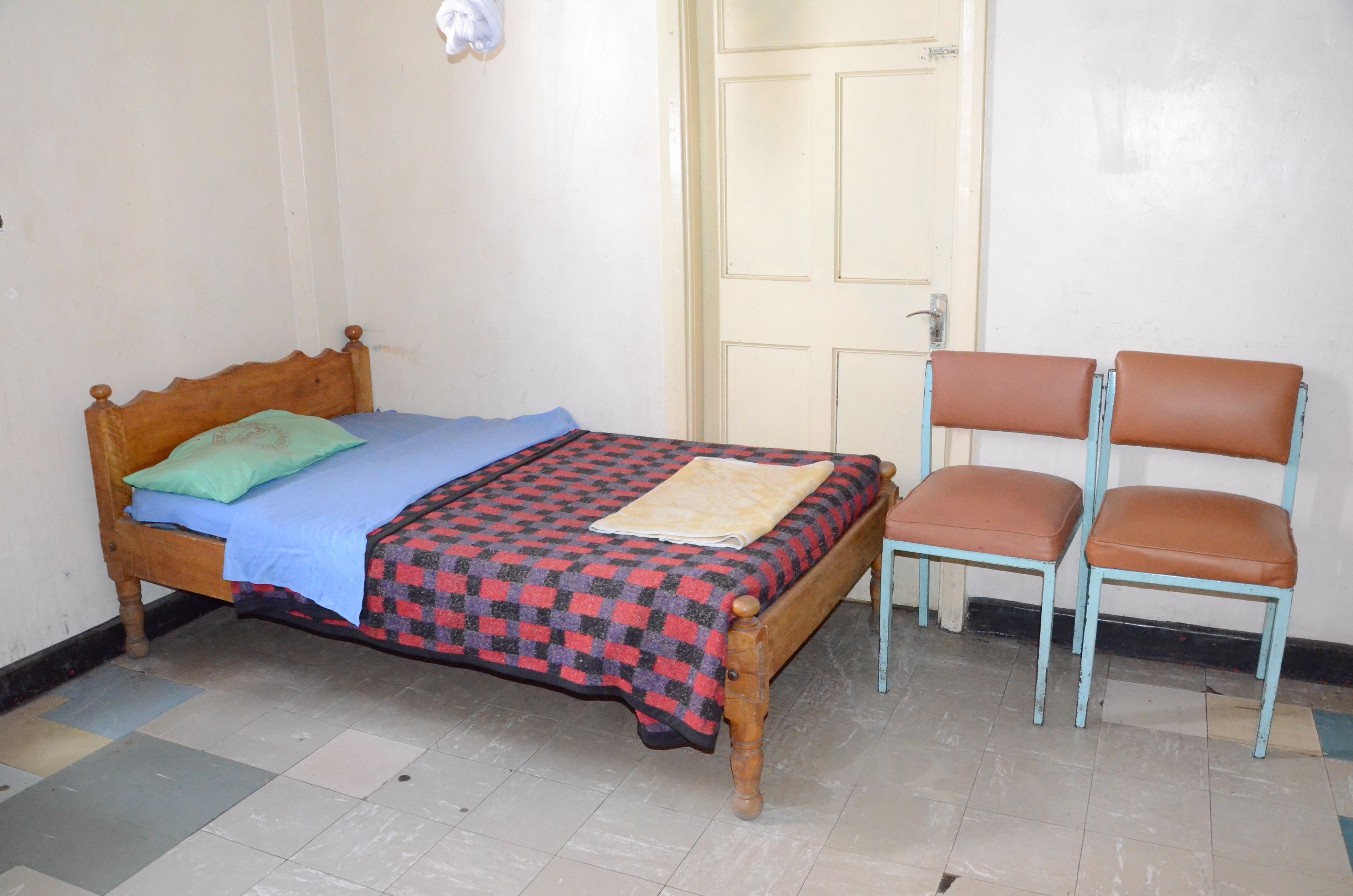 A single bed with a checkered bedspread and pillows, a chair with a cushion, and a door in the background.