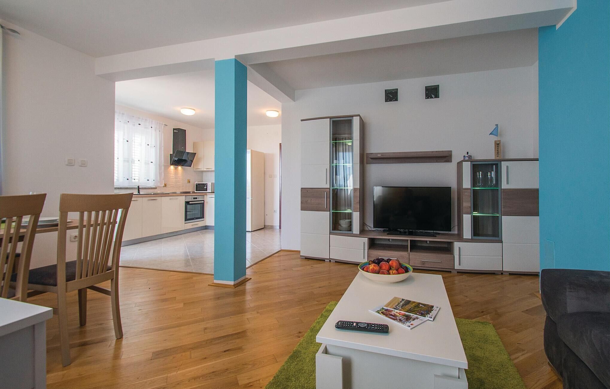 Een moderne woonkamer met een flatscreen televisie, een salontafel met een schaal fruit, en een eethoek met stoelen en een tafel.