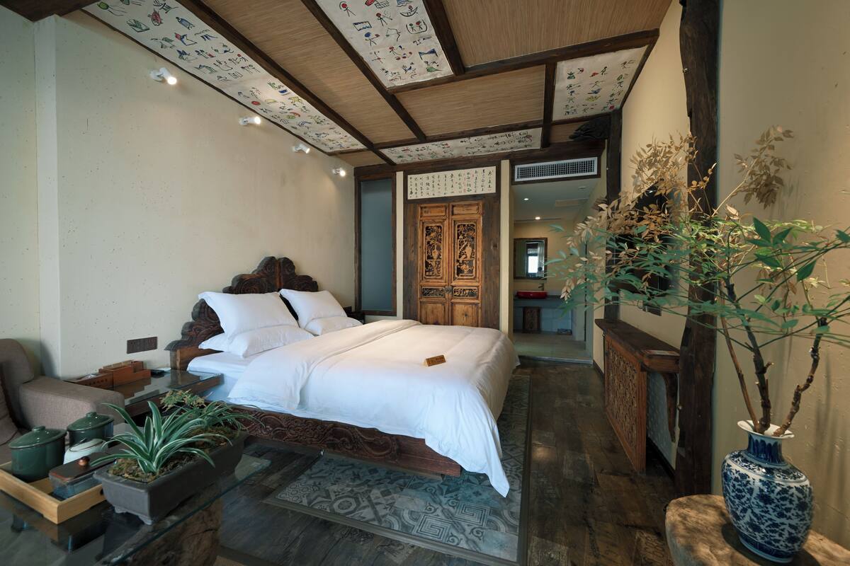 A traditional room with a bed, wooden furniture, a vase with plants, and a decorative ceiling.