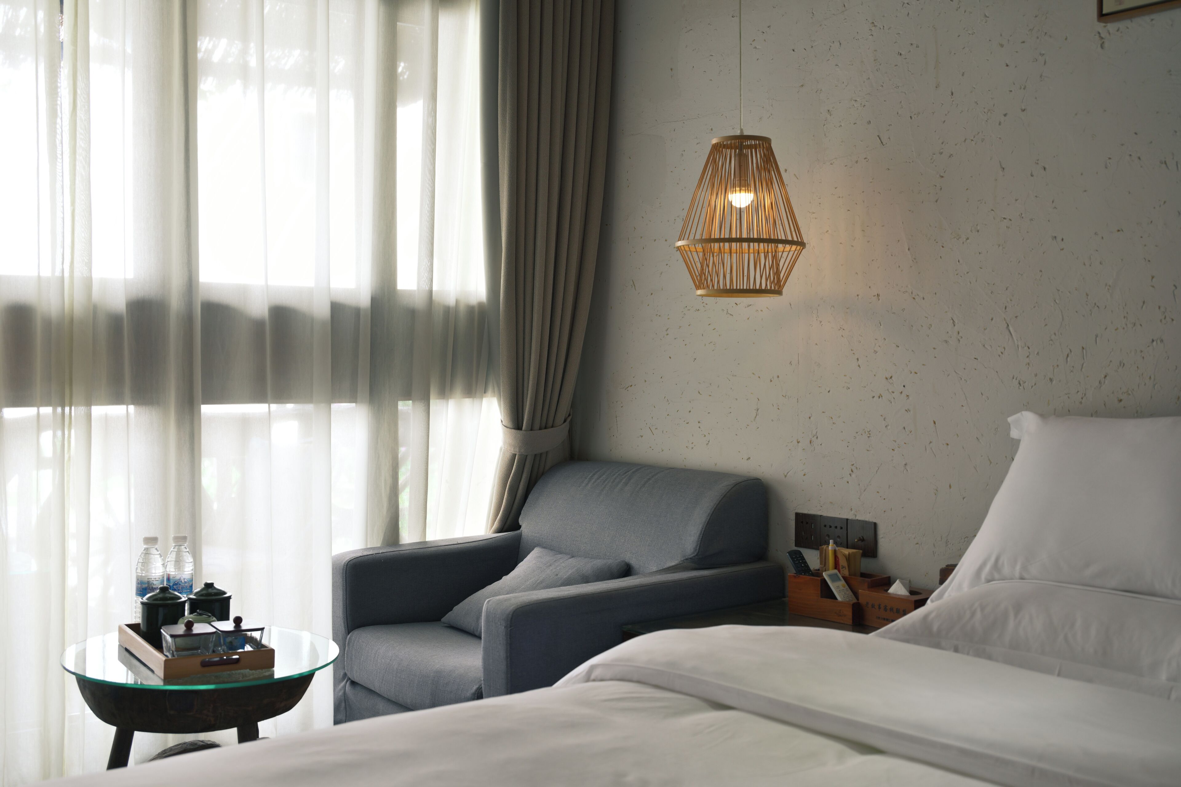 A hotel room with a bed, a chair, a glass table with a tray of items, a hanging light fixture, and a window with curtains.