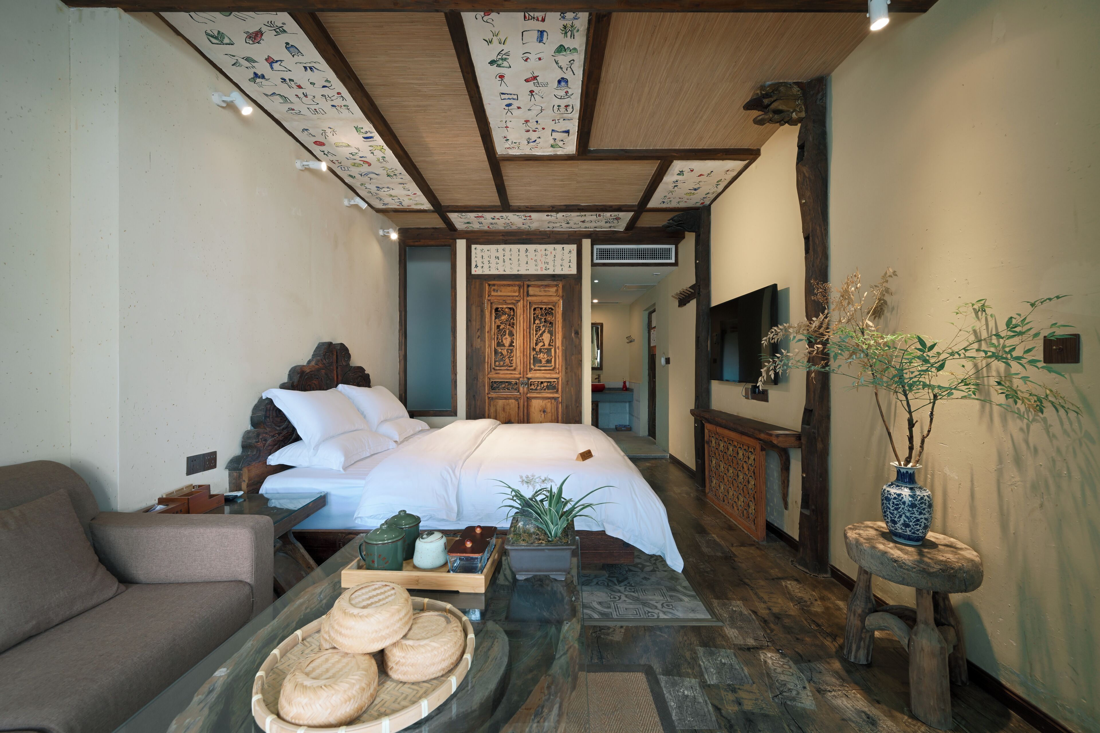 A traditional room with a bed, a sofa, a small table with a tray of bread, a vase with a plant, and a wooden cabinet.