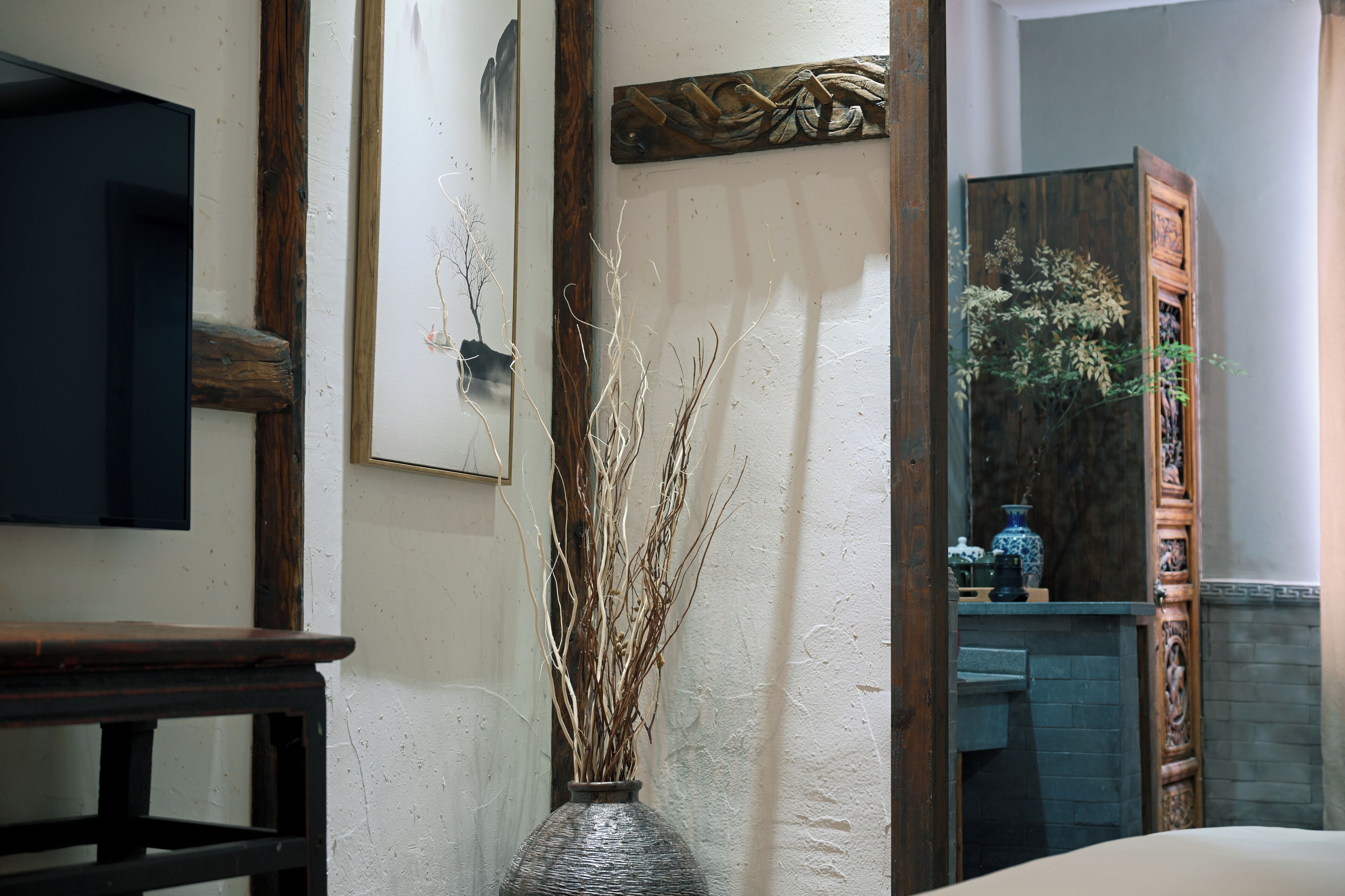 A room with a wooden table, a vase with dried branches, and a mirror reflecting a cabinet with decorative items.