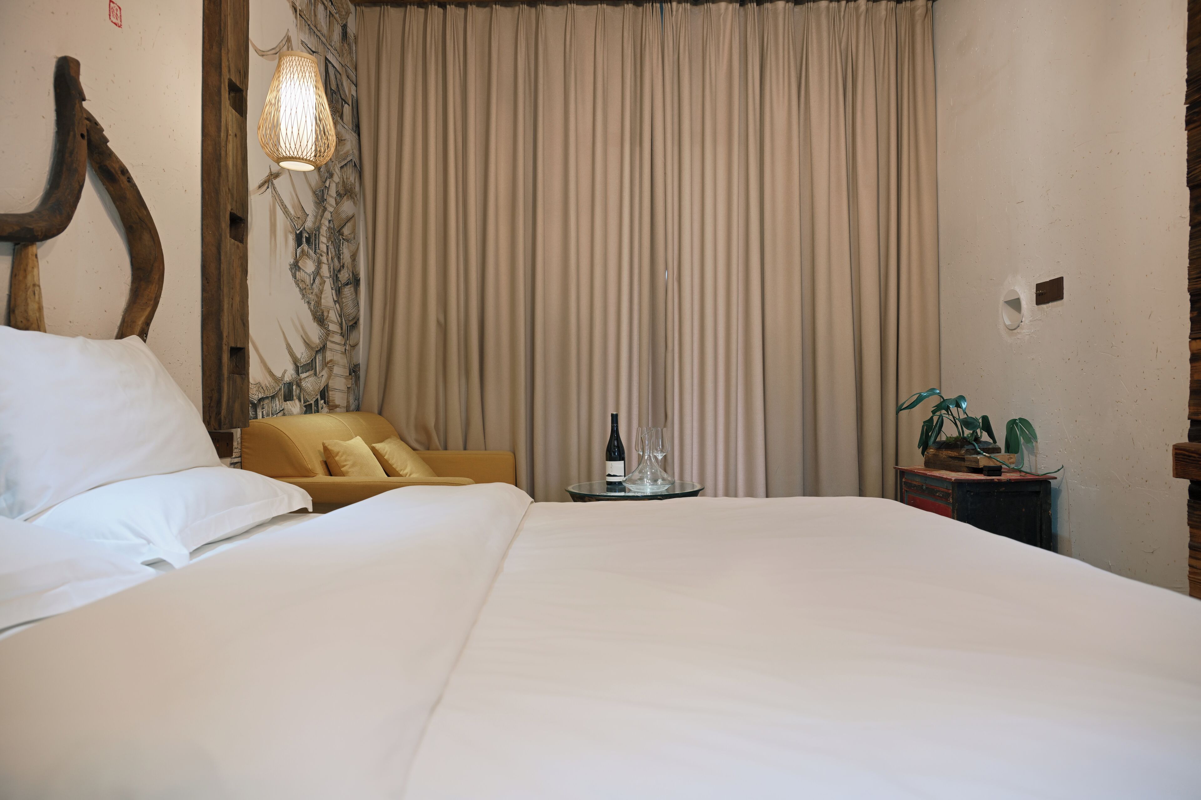 Quarto de hotel com uma cama grande, um sofá pequeno, uma mesa com uma garrafa e copos, e uma planta.