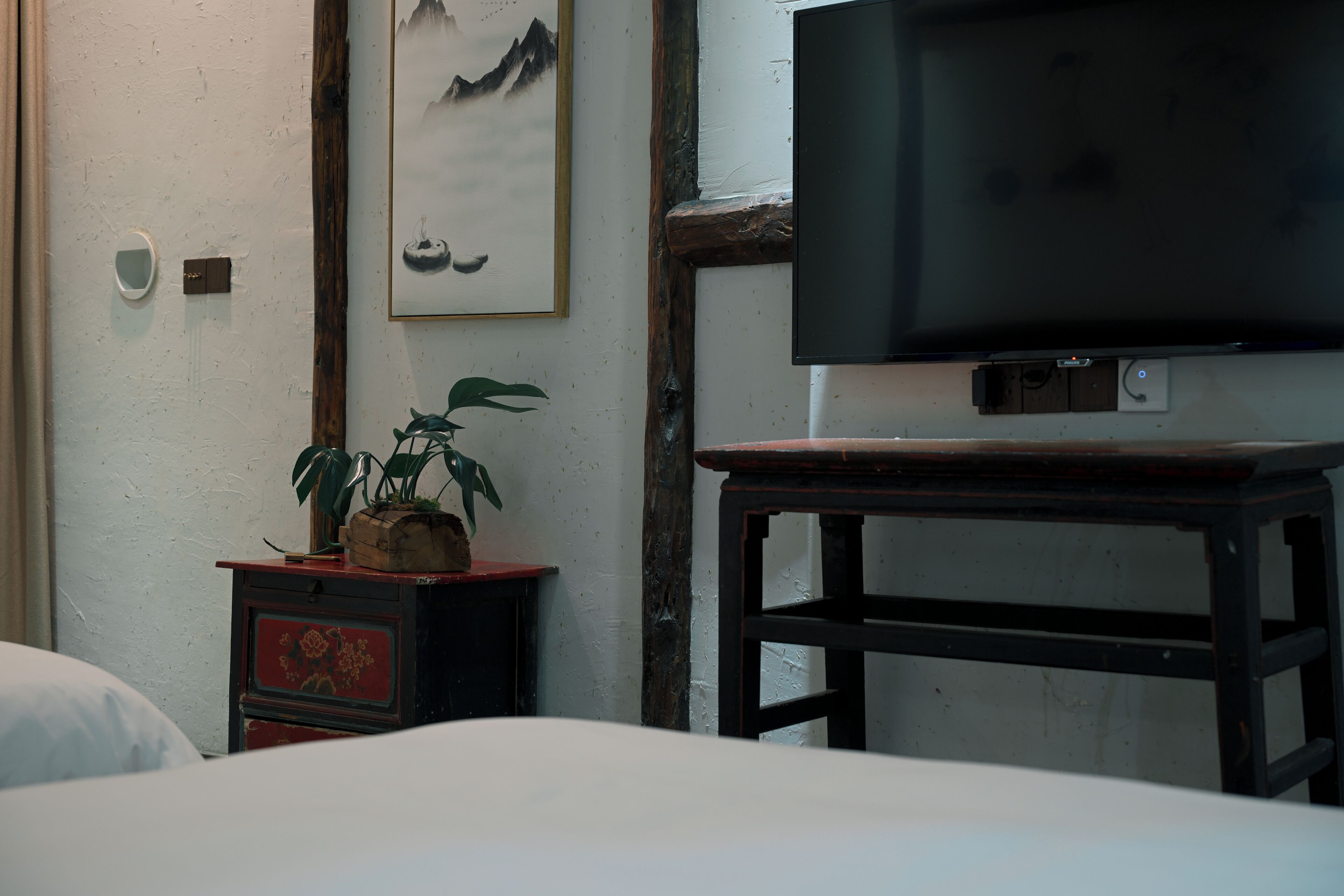 Una habitación con una cama, una mesita con una planta, un televisor en la pared y un armario de madera.