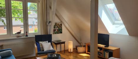 A cozy room with a sloped ceiling, a sofa, a coffee table, a bookshelf, and a window with a view of greenery.
